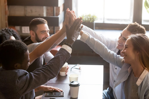 Multiracial friends giving high five, sitting together at coffee table, enjoying drinks in cozy cafe, diverse millennial workers joining hands showing unity at meeting in coffee shop. Close up view